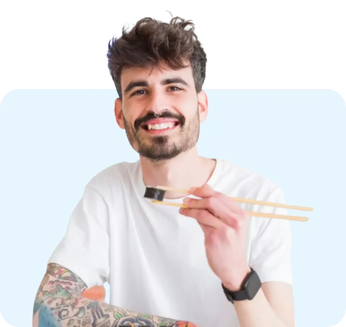 Smiling man holding sushi with chopsticks against a light blue background.